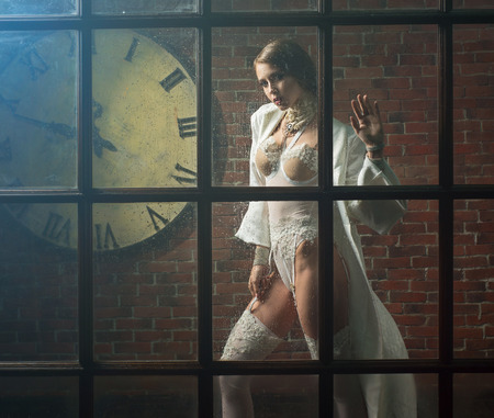 Slim woman in white lingerie is photographed through a window near a brick wallの写真素材