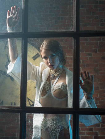 Slim woman in white lingerie is photographed through a window near a brick wallの写真素材