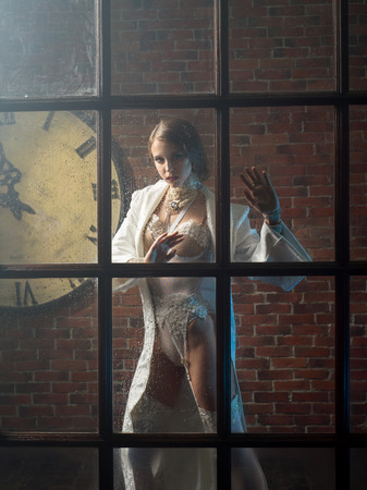 Beautiful woman in white lingerie is photographed through a window near a brick wallの写真素材