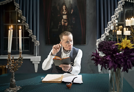 Imposing man sitting at a desk and looks through a magnifier in bookの写真素材