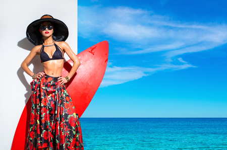 Beautiful woman in a black hat, bra and bright skirt on the background seascapeの写真素材