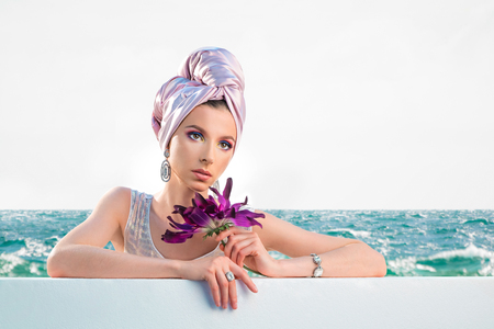 Beautiful woman in shiny white swimsuit and in headscarf with flower on seaの写真素材
