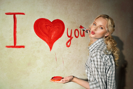 Young woman in plaid shirt with paintbrush is drawing a heart and inscription of love on the wallの写真素材