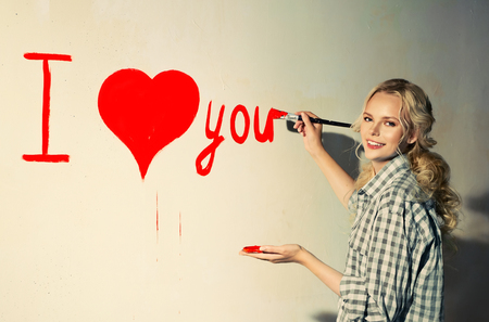 Young woman in plaid shirt with paintbrush is drawing a heart and inscription of love on the wallの写真素材