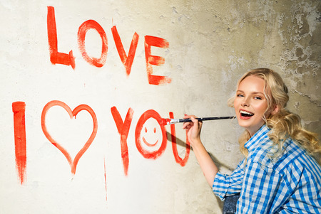 Young woman in plaid shirt with paintbrush is drawing a heart and inscription of love on the wallの写真素材