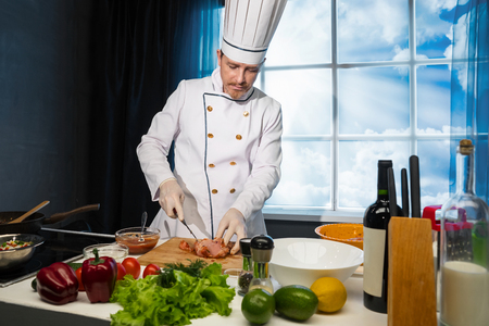 Cook in white uniform cuts chicken with knife on wooden board in the kitchenの写真素材