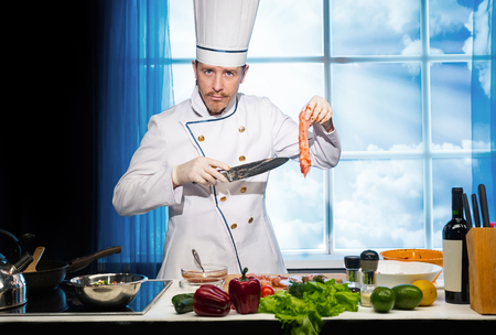 Cook in white uniform cuts chicken with knife on wooden board in the kitchenの写真素材