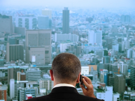 Businessman discussing to the mobile phone in his office while is looking through the window to the city.Focus on the businessman,the city aspect is blured.の写真素材