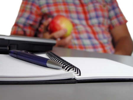A schoolboy holding an apple at the desk. Focus on the notebook and ballpen.