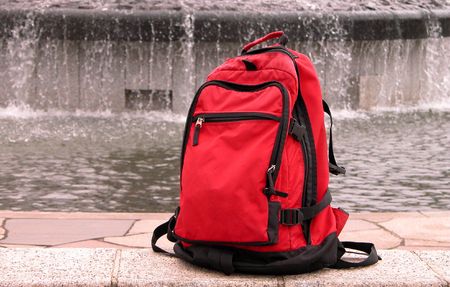 A red backpack in a park. As background is an artesian well.Images suggesting travel......focus on...