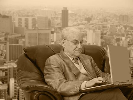 Senior businessman working on his laptop in front of the office window in a big city. The sepia...