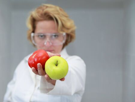 Image of a female researcher offering a tomatoe and an apple to suggest the ideea that healthy eating is recommended also by scientists.Specific lighting for a classical research laboratory.の写真素材