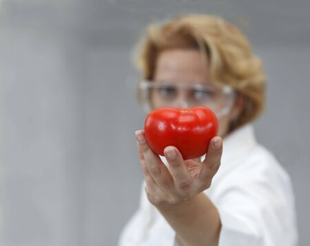 Image of a female researcher offering a tomato and an apple to suggest the idea that healthy eating is recommended also by scientists.Specific lighting for a classical research laboratory.の写真素材