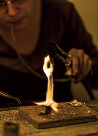Female jeweller blowing a torch to melt metal for making a brooch in her workshop.の写真素材