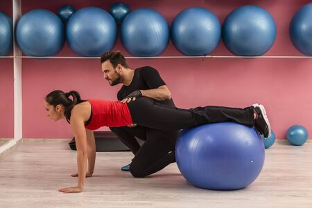 Young woman helped by her coach is doing exercises with a fitness ball  in a gym.