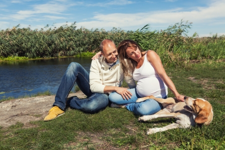 Family portrait - pregnant woman and her husband are sitting in a park next to their happy dogの写真素材