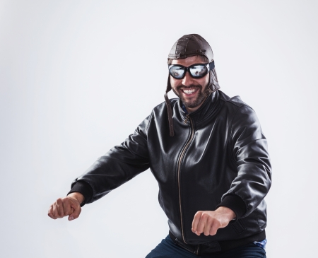 Funny young man, posing as a motorcyclist, is wearing black leather jacket, vintage cap and glasses - isolated on whiteの写真素材