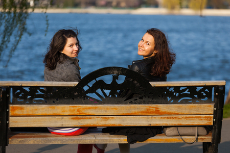Closeup of two smiling woman sitting on a bench near a lake and looking behind to the cameraの写真素材