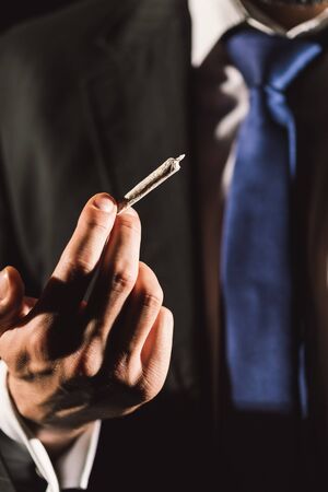 Businessman holding a joint in his hand. Focus on the fingers and man in blur in the background.の写真素材