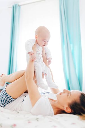 Young mother holding her baby boy on knees and lying in bedの写真素材