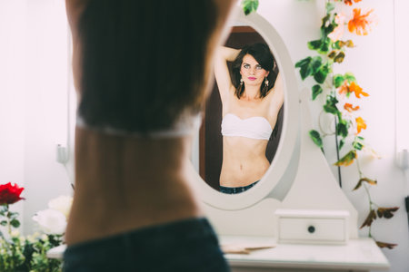 Beautiful female posing in front of the mirror, in a white bra and jeans, in a very seductive wayの写真素材