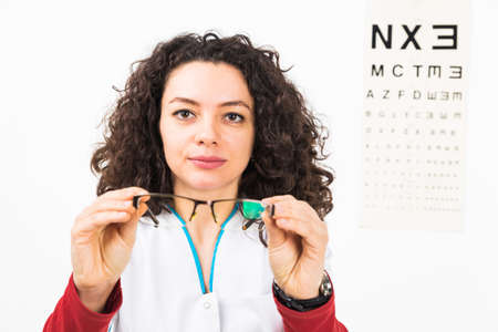Closeup of a serious woman doctor holding eyeglasses with eye chart behind - isolated on white.の写真素材