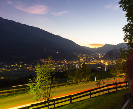 Car stop lights create a continuous line on the road at sunset. Long exposure technique. Lighted city in an Austrian valley in the background.の写真素材