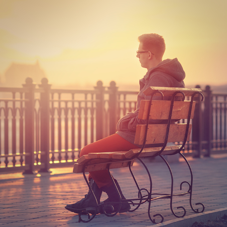 Lonely Man Sitting on a Bench during Sunset. Young Man Sitting on Wooden Bench. Sunset Male Outdoor Backgroundの写真素材