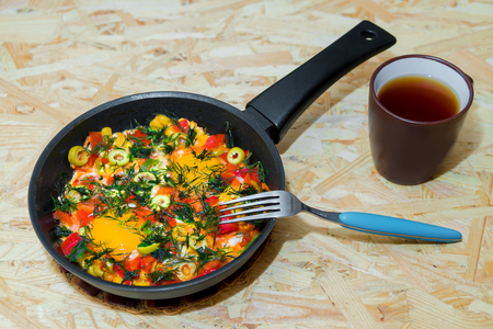 Eggs in a frying pan with pepper, olives, greens, fork and a cup of teaの写真素材