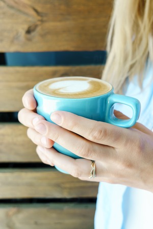 Closeup view on blue cup of cappuccino holding by young girl in jeans wear. Wooden background.の写真素材
