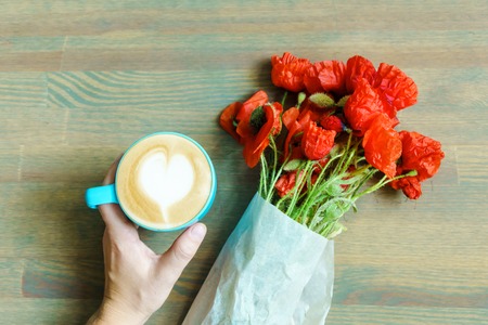 Spring concept. Woman hand with heart froth cappuccino and poppy flower on wooden table.の写真素材