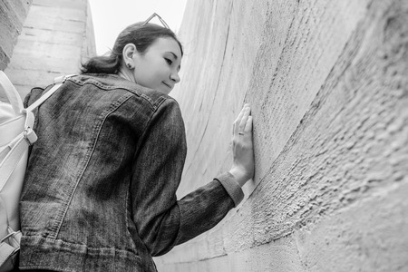Attractive young woman stands between concrete walls. Caucasian girl in casual jeans and flower dress looks back. Pretty fashion woman in outdoor space.の写真素材