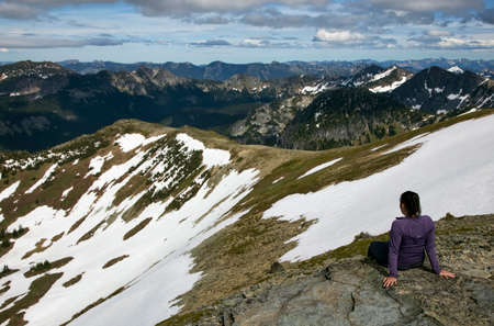 A female hiker stops to rest and enjoy the view at the top of a partially snowy mountain.の写真素材