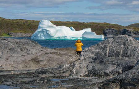 A female tourist in a bright yellow coat photographs an iceberg near Noddy Bay, Newfoundland.の写真素材