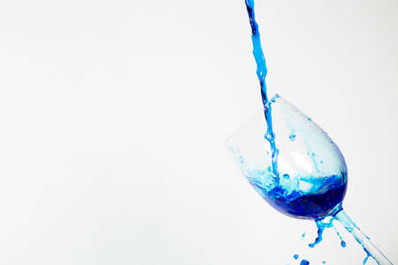 A stream of poured bright blue liquid splashes into a tilted clear wine glass, on a clean white background.の写真素材