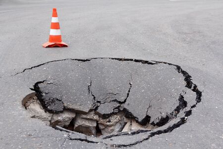 Deep sinkhole on street city and orange traffic cone. Dangerous hole in asphalt highway. Road with cracks and pit. Bad construction. Damaged surface road collapse and fallen. Broken infrastructure.の写真素材