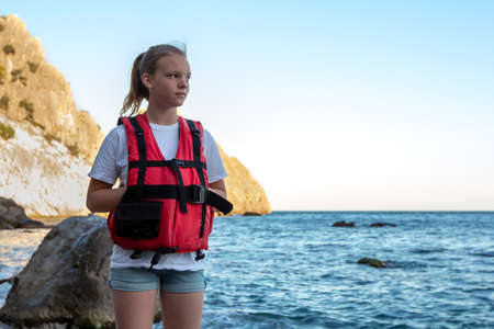 Young girl in red life jacket stands on beach with rock and sea. Female lifeguard on duty of coast. Water safety conceptの写真素材