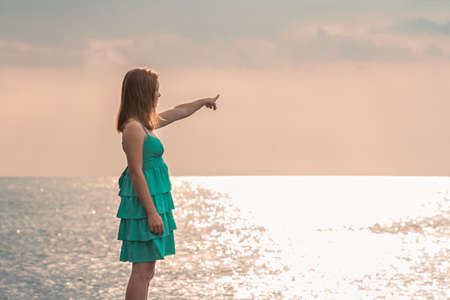 Young girl in blue dress pointing to sea. Beautiful blonde with long hair stands on ocean in summer and points hand into distance.の写真素材