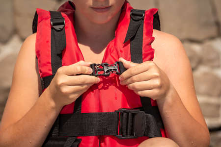 Young girl putting on red life jacket on the beach on a summer sunny day. Female hands fastens plastic buckle clasp of life vest. Safety during water sportsの写真素材