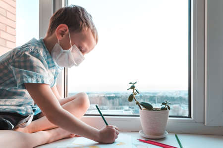 Kid sits at home by window in medical mask and draws with pencils during quarantine and epidemic.の写真素材