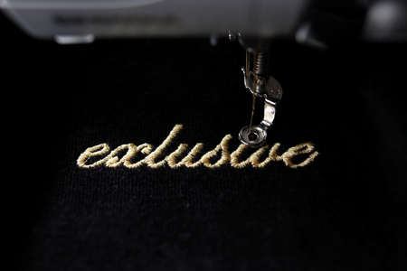 embroidery of gold lettering "exclusive" on black velvetely fabric with embroidery machine - front view with part of machine - background and foreground blanked out blurryの写真素材