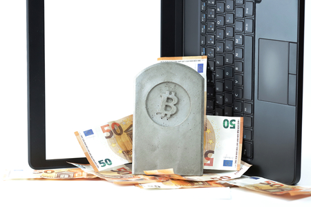 stone monument/tombstone with bitcoin symbol standing on a pile of banknotes in front of a flipped black notebook on white background - copy space for text on displayの写真素材