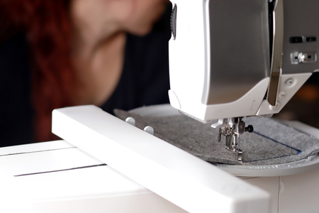 View on machine head of a modern sewing machine with embroidery unit stitching a blue frame on grey felt - background with redhead woman blanked out blurryの写真素材