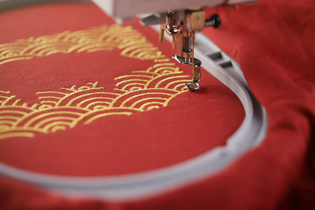 Oblique view on embroidery of traditional shell pattern framing pig outline with gold on red fabric by modern embroidery machine - chinese new year concept - view on stitching area with needle moving downの写真素材