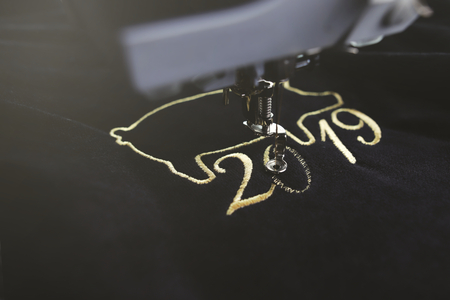 Embroidery machine stitching 2019 chinese new year motive with precious gold yarn on black velvet in dark misty light mood - oblique view with blurred backgroundの写真素材