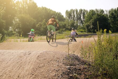 mother and two daughters riding bike together on a bicycle dirt track in bright summer light - family lifestyle outdoor activity concept - focus on hill tip in foreground - rider blanked out blurry in backgroundの写真素材