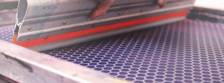 Close up on a hand-guided screen printing squeegee shoving silver paint over a silk-screen frame - handicraft and exclusive fashion concept - background blanked out blurryの写真素材