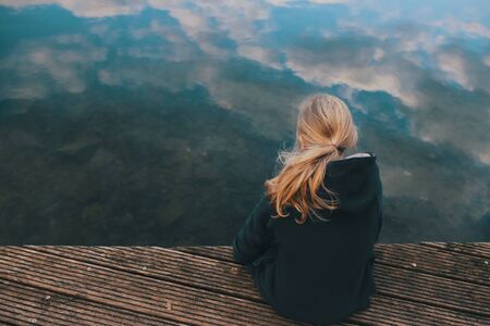 young girl with long blonde hair is sitting on a wooden stage looking in clear shallow water with reflecting cloudy sky in a thoughtful pose - coming of age, sadness and sorrow conceptの写真素材