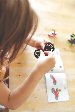 blonde haired young girl assembles gears with her little fingers to build a toy tractor from  bricks while looking a the assembly instruction - bright light mood with blurred background - portrait orientationの写真素材