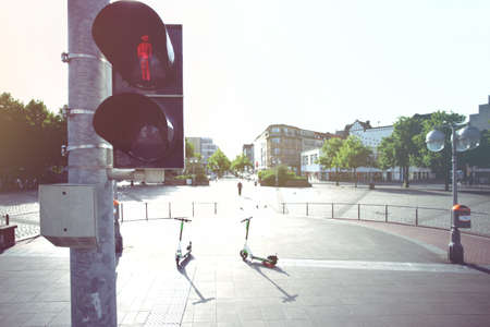 pedestrian traffic light shows a red standing character graphic in front of a wide inner city square with electric scooters, pedestrian and city buildings in bright toned lightの写真素材